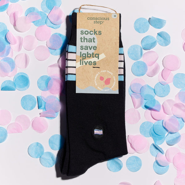 a pair of black socks in cardboard packaging that reads "socks that save lgbtq lives"with a small embroidered trans pride flag patch on the ankle and 5 stripes of blue, pink and white near the cuff symbolizing the trans pride colors