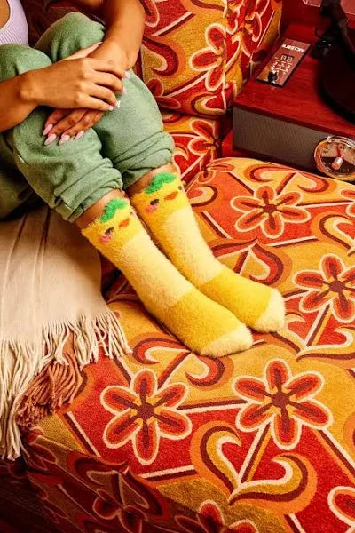 A model shown from the knees down sits on a funky sofa. They wear fuzzy yellow crew socks with duck faces and a leaf on the head.