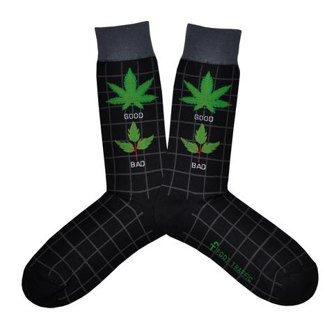 Shown in a flatlay, a pair of Foot Traffic black cotton men's crew socks with a pot leaf titled in text “Good Weed” and a poison oak plant titled in text “Bad Weed”