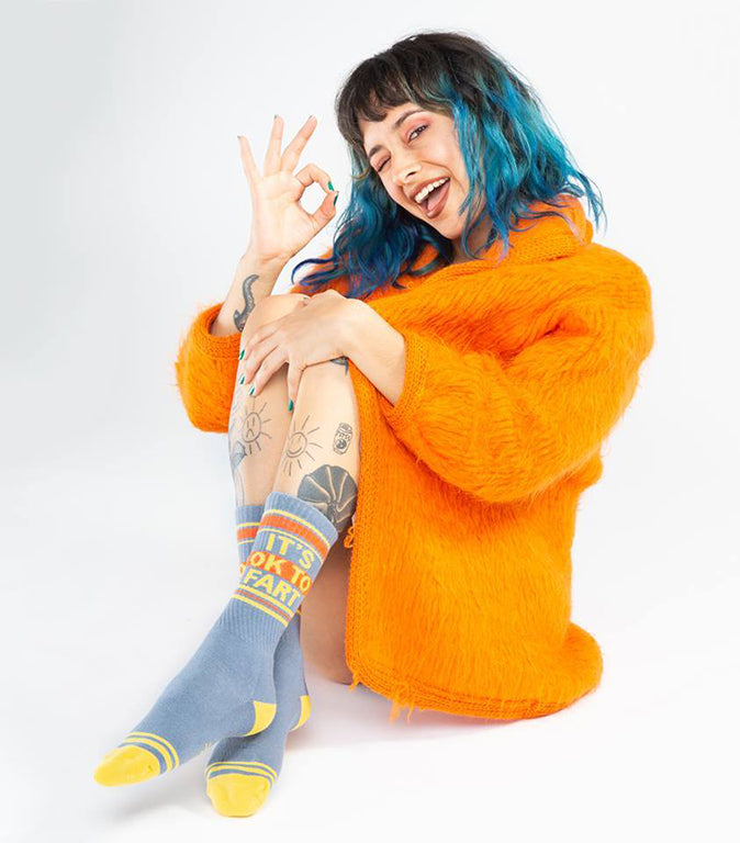 Shown on a tattooed feminine model, a pair of grey cotton Gumball Poodle brand unisex crew socks with yellow and orange stripes on the cuff and yellow striped heel and toe. These socks feature the phrase, 