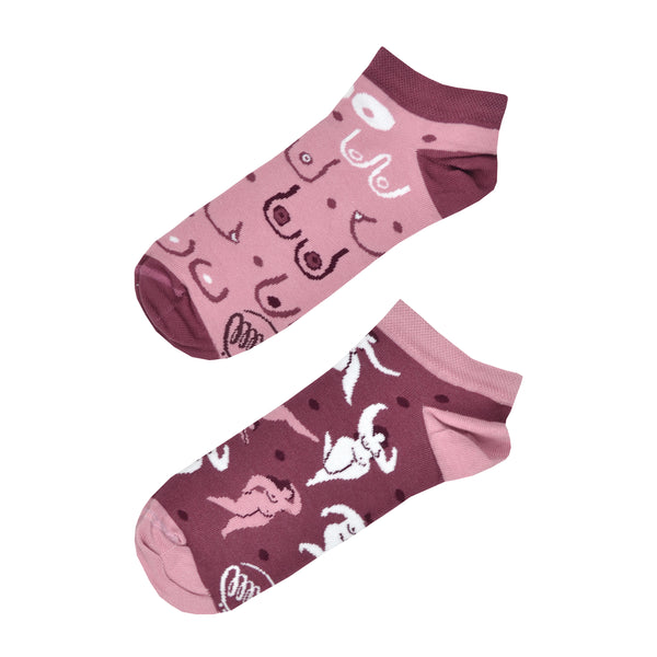 Shown in a flatlay, a pair of mismatched Many Mornings brand unisex cotton ankle socks. One sock has a dark pink heel/cuff/toe with a light pink base and cartoon images of different breast shapes. The second sock has light pink heel/cuff/toe and a darker pink base features an all over design of curvaceous nude feminine bodies.