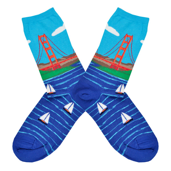 These blue cotton women's crew socks by the brand Socksmtih feature the iconic landmark of San Francisco, the Golden Gate Bridge, on a clear sunny day towering above the beautiful bay filled with sailboats.