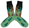 Shown in a flatlay, a pair of Sock It To Me green cotton men's crew socks with black starry sky and various sasquatches enjoying a campfire (playing banjo, roasting marshmallows) among a pine forest