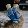 A model sits on a rock while wearing denim-blue socks with tree patterns and a yellow sky at the ankles
