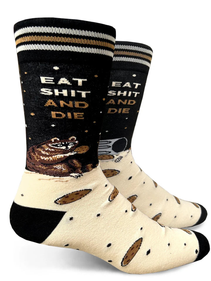 A white background shows a pair of crew socks on foot forms. The feet of the socks are beige with black toes & heels and a pattern of small black dots and large chocolate chip cookies. The ankle is black with several brown and white dots that look like stars. On the outer edge of the ankle a pudgy raccoon sits on its haunches with a cookie under beige and brown text that reads 