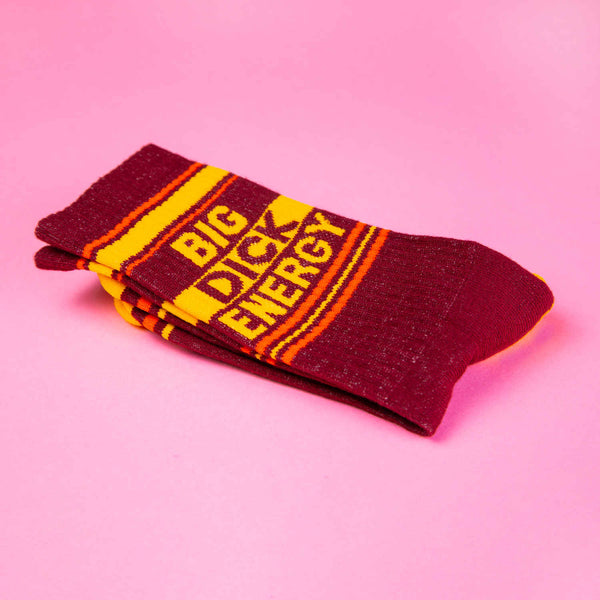 burgundy socks laid flat with a yellow toe and heel and orange stripes with bold text that reads BIG DICK ENERGY folded in half and laying on a pink background