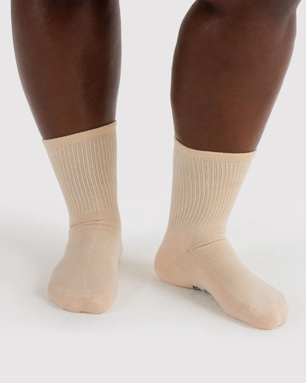 A close-up of a person's lower legs and feet wearing ribbed crew socks in a light beige or nude color, set against a plain light gray studio background.