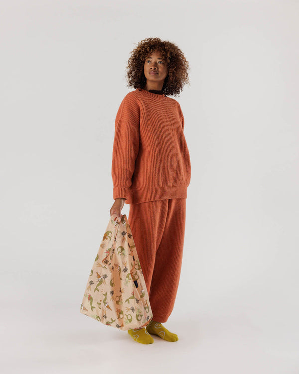 a Black woman wearing a matching orange sweater set and pants holding a light pink reusable shopping back with a print of illustrations of rabbit mermaids and small bunches of carrots