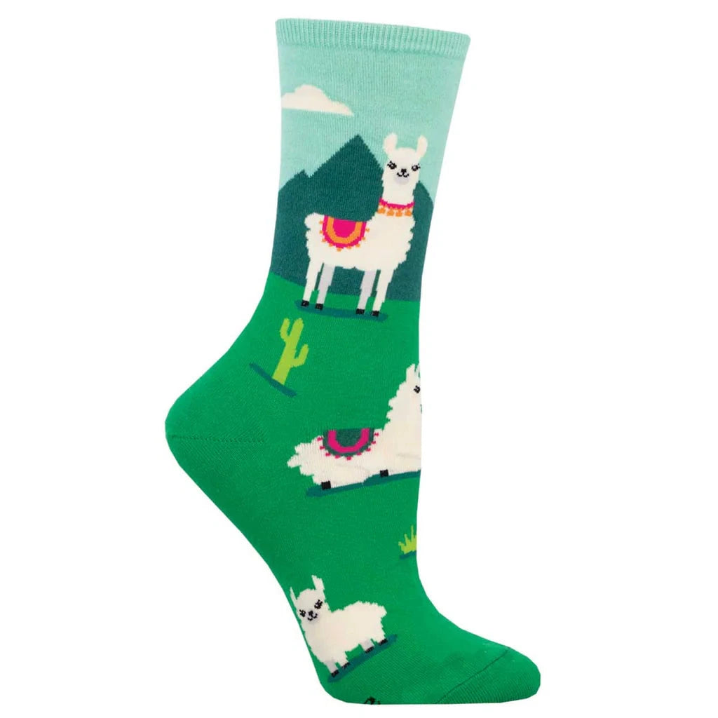 Foot form wears green crew sock featuring white llamas in pink and gold necklaces and saddles, cactuses and a cloudy mountain backdrop