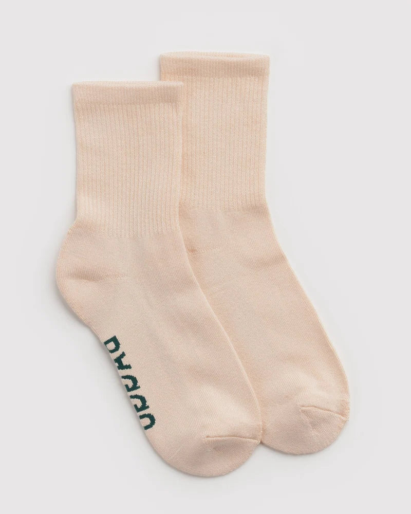 A pair of ribbed crew socks in a light peach-beige color, with the brand name 'BAGGU' printed vertically in dark green on the side of one sock, set against a plain light gray background.