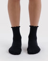 Black ribbed ankle socks with a lettuce edge, shown on a person's feet against a light gray background.