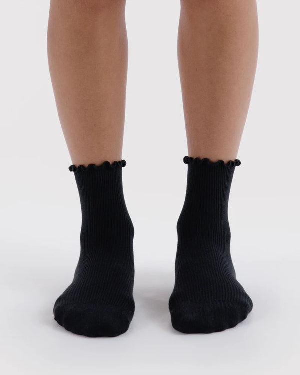 Black ribbed ankle socks with a lettuce edge, shown on a person's feet against a light gray background.