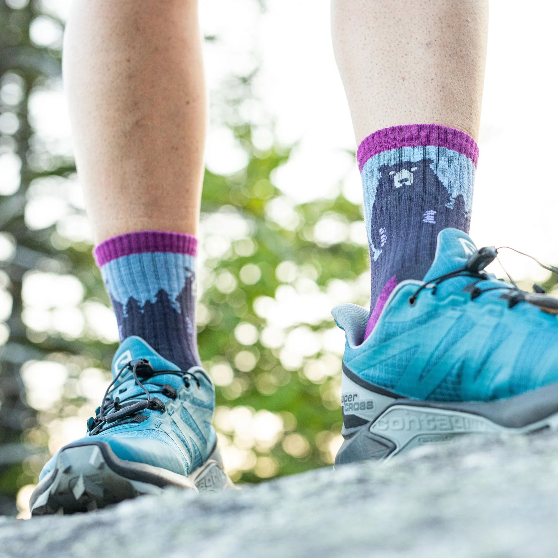 Model wears hiking shoes and blue/purple socks featuring a bear and a treeline