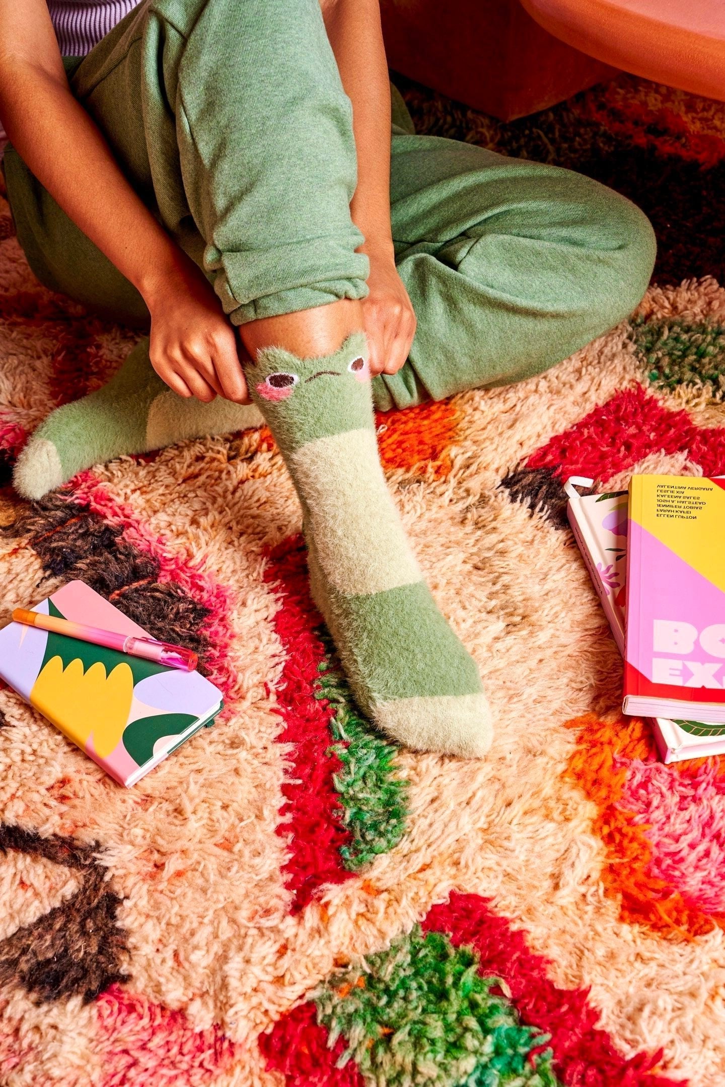 A model is shown sitting from the knee down. They sit on a funky carpet while wearing fuzzy green crew socks with cute frog faces at the top.