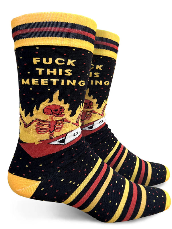 White background with two black crew socks on foot forms featuring yellow and dark red stripes around the ankle and foot, small red and yellow speckles and a red skeleton sitting at a red desk in front of a white laptop. The skeleton is on fire, and yellow text above it reads "FUCK THIS MEETING."