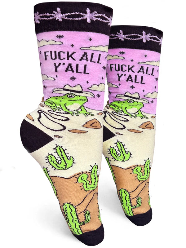 White background with two pink crew socks on foot forms with black barbed wire pattern around the top, beige and tan sand on foot, multiple green cactuses, and a green frog wearing a beige cowboy hat holding a whip. Black text reads "FUCK ALL YALL."