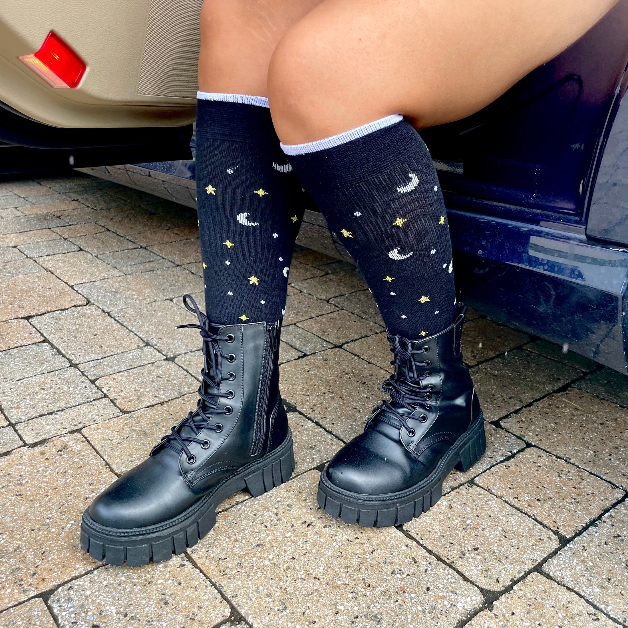 a cropped photo of a pair of tanned legs wearing black knee high socks with a heathered grey toe and heel and pattern of petite white and gold stars and crescent moons and chunky black boots