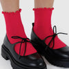 Bright red ribbed crew socks with ruffled tops worn with black Mary Jane-style platform shoes featuring black laces, shown on a person's feet against a light gray background.