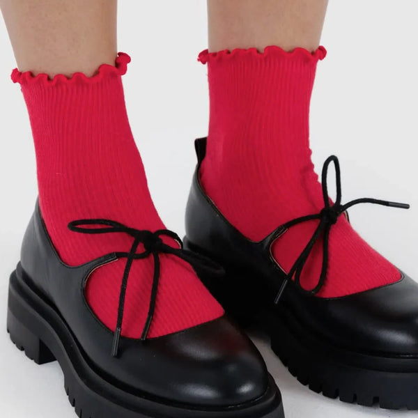 Bright red ribbed crew socks with ruffled tops worn with black Mary Jane-style platform shoes featuring black laces, shown on a person's feet against a light gray background.