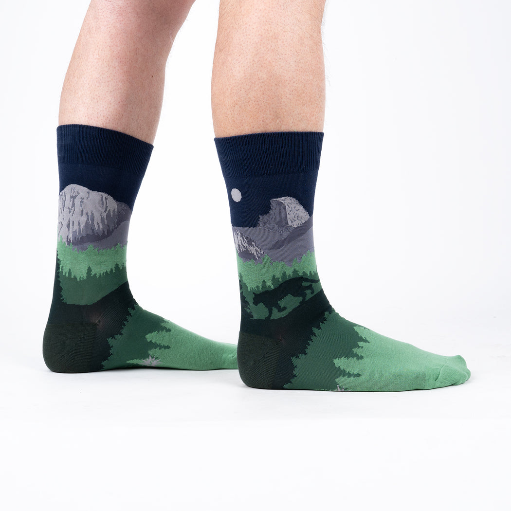 A white background shows a pair of legs wearing crew socks that portray a dark blue night sky and Yosemite's half dome on the legs and three different shades of green foliage. The silhouette of a cougar walks along the outside ankle.