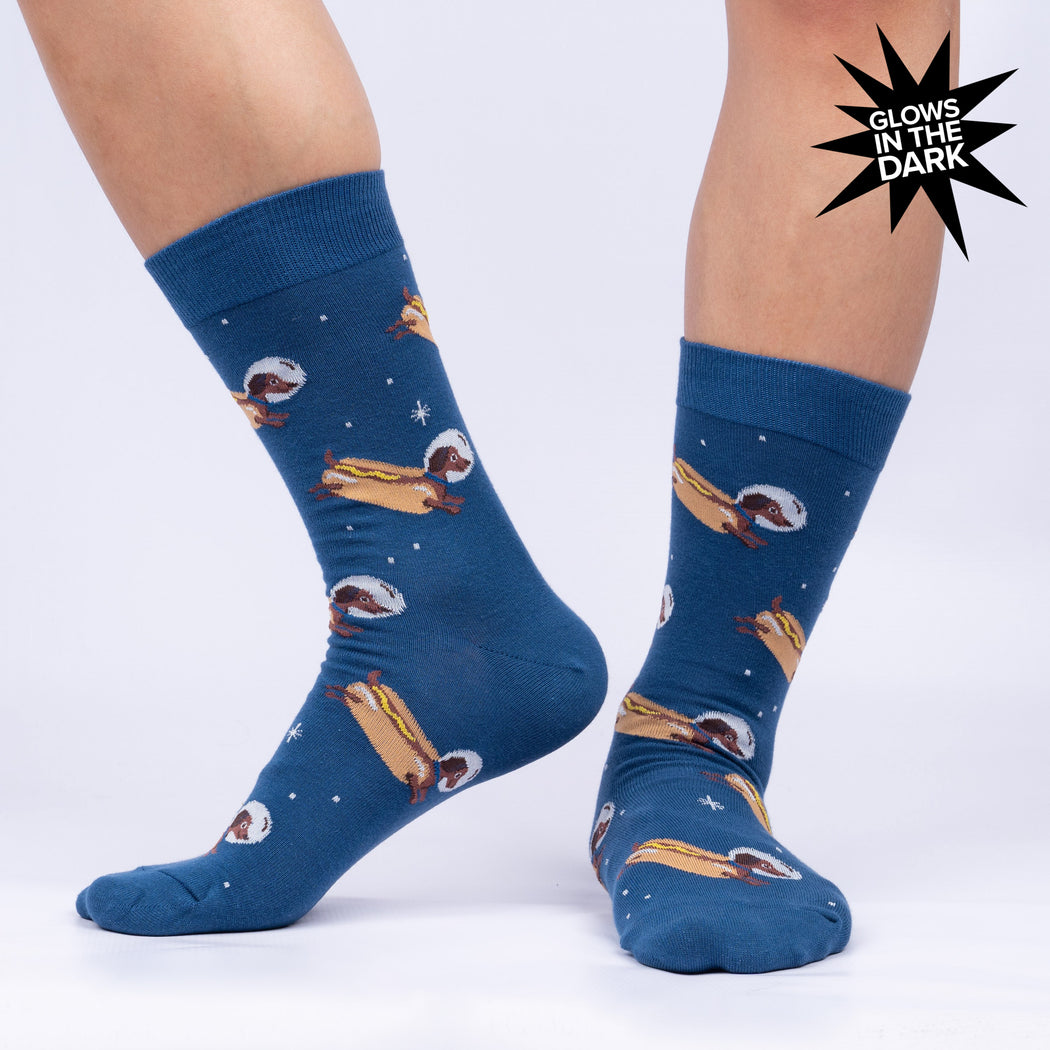 We see two feet dressed in dark blue socks with small white stars and dachshunds with the bodies of hot dogs wearing astronaut helmets. One foot is shown from the front, the other the side. A black blurb that reads 