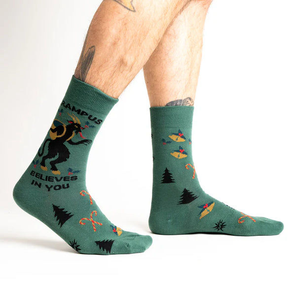 Model wears teal socks with Krampus and holiday motifs. Text reads "KRAMPUS BELIEVES IN YOU"