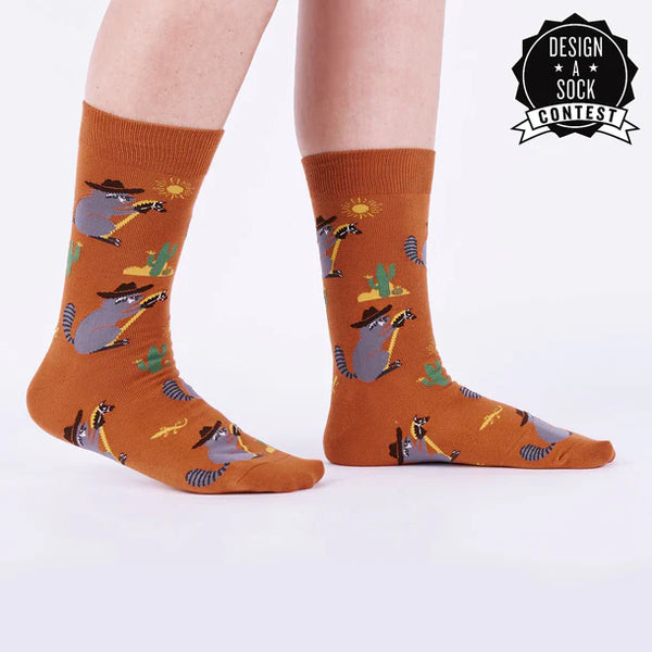 Model shown from knees down wears brown socks featuring raccoons wearing cowboy hats riding hobby horses through a desert scene