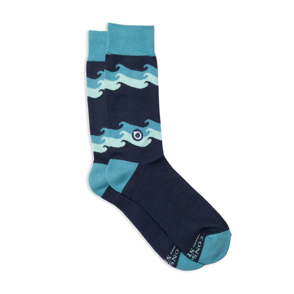 Shown in a flatlay, a pair of unisex crew socks in navy blue with a teal heel, toe, and, cuff. The leg of the sock features blue ocean wave patterns.