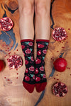 A pair of black crew socks with a red and green pomegranate pattern, worn by a person whose legs are shown from the calves down, styled on a wooden surface surrounded by whole and sliced pomegranates.
