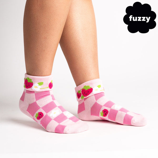 A white background holds a pair of legs wearing hot-and-light pink checkerboard socks with fuzzy white daisies and smiling strawberries on some of the squares. The ankle is turned down to reveal more strawberries and daisies ringing the inside.