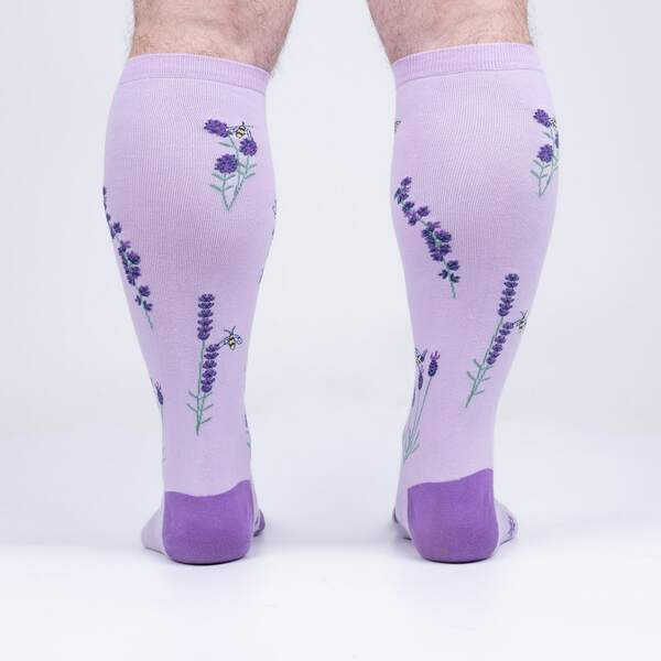 a pair of hairy legs, seen from the back, wearing a pair of light purple knee high socks with a darker purple toe and heel covered in small purple flowers, green stems and small bees