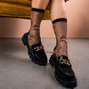 Model sits on couch wearing chunky black shoes and sheer black socks with yellow & orange beetle designs