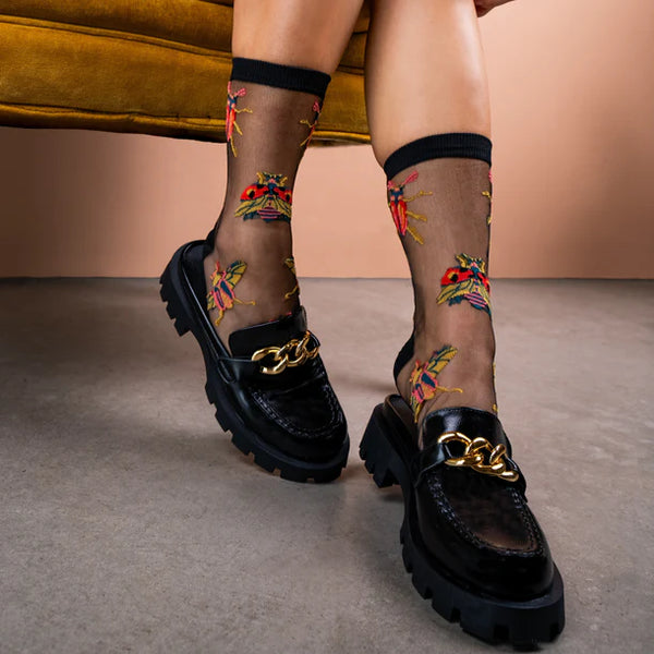 Model sits on couch wearing chunky black shoes and sheer black socks with yellow & orange beetle designs
