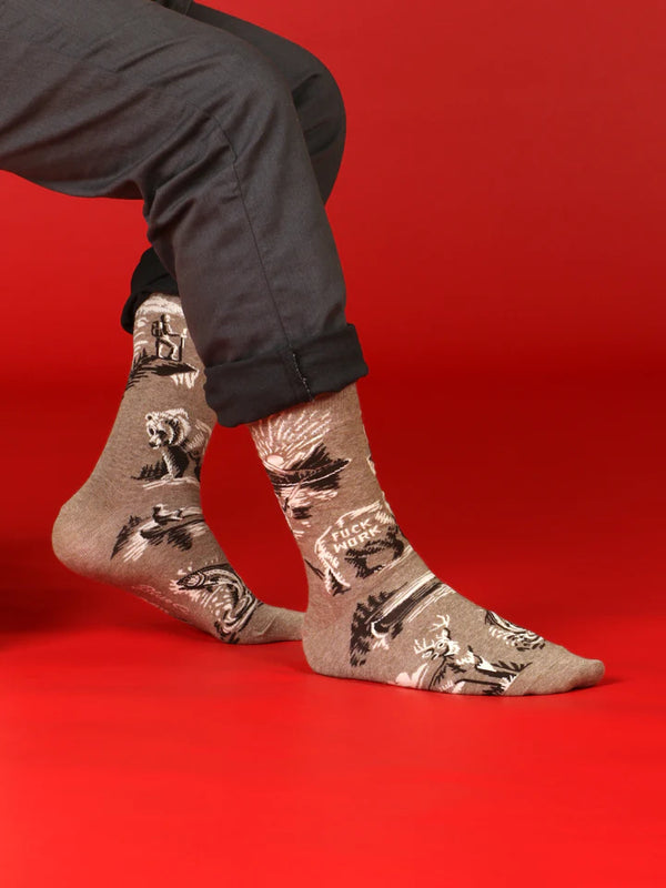 Model shown from legs down wearing light brown crew socks with designs of a fisherman at a mountain lake sunset, deer, canoes, and a bear with text that reads "FUCK WORK" on its side