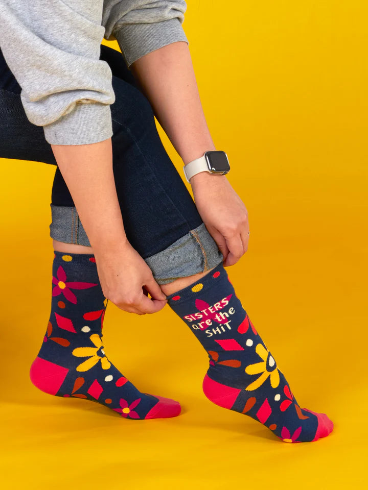 Yellow backround, model shown from the waist down adjusting a pair of navy blue crew socks with floral and geometric designs in hot pink, magenta, brown and gold with beige text that reads 