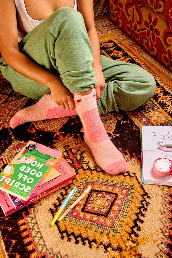 A model shown from the waist down sits on a funky carpet surrounded by books and journals. They wear a pair of pink fuzzy crew socks with blushing frog faces at the top.