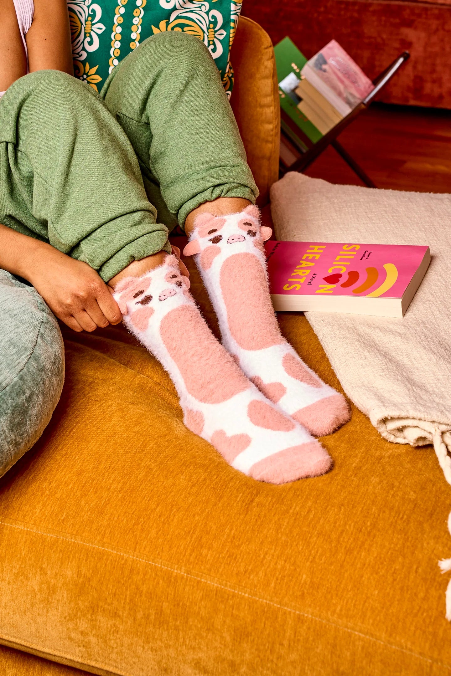 A model is whon from the knees down sitting in a warm living room scene. They wear fuzzy crew socks that look like pink-and-white 3d cows. 