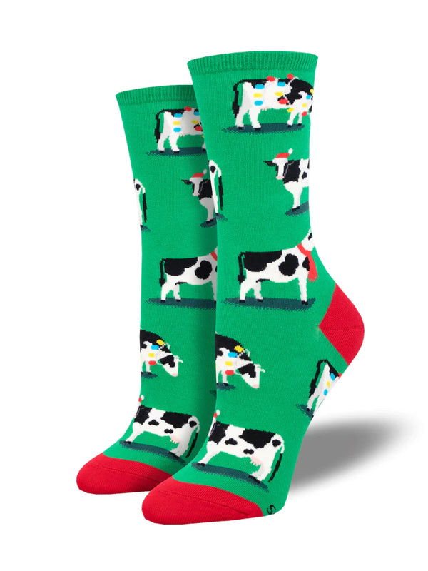 Shown on leg forms, a pair of women's crew socks in green with a red heel and toe. These socks have an all over motif of black and white cows in holiday gear. Wrapped in lights and wearing santa hats. 