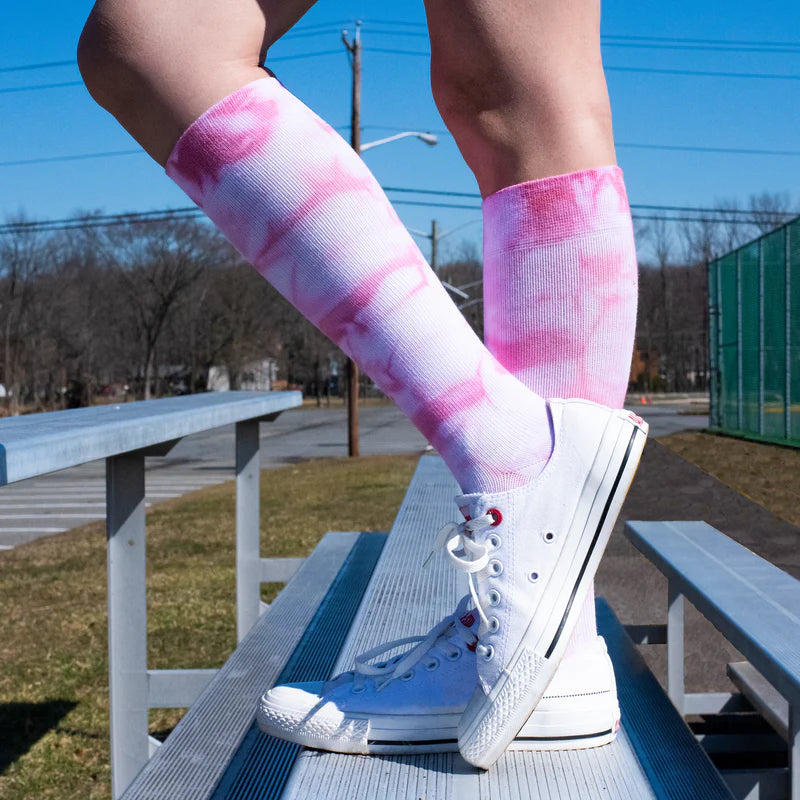a cropped photo of a woman standing on metal bleachers outdoors wearing white sneakers and pink and white tie dye knee high socks