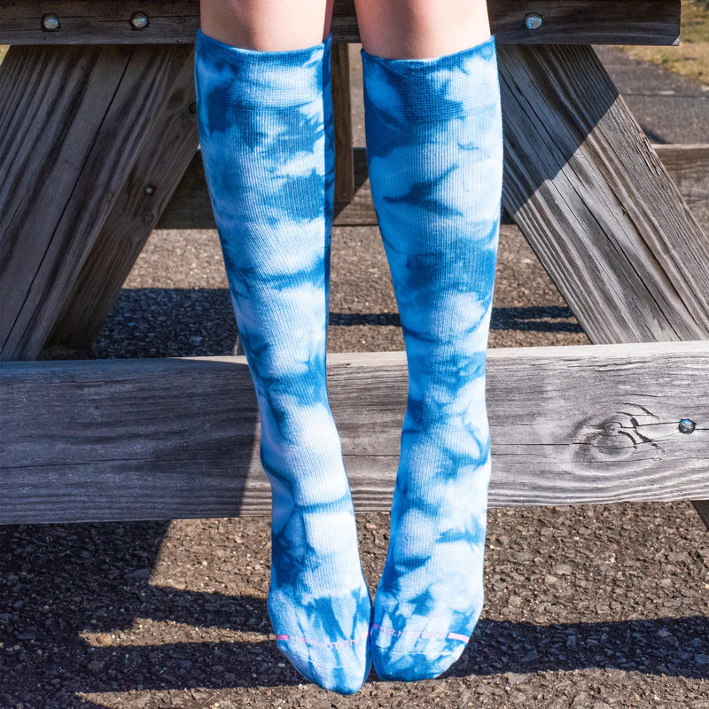 a photo of a woman's legs dangling off the side of a wooden picnic table, wearing a pair of tie dye blue and white knee high socks
