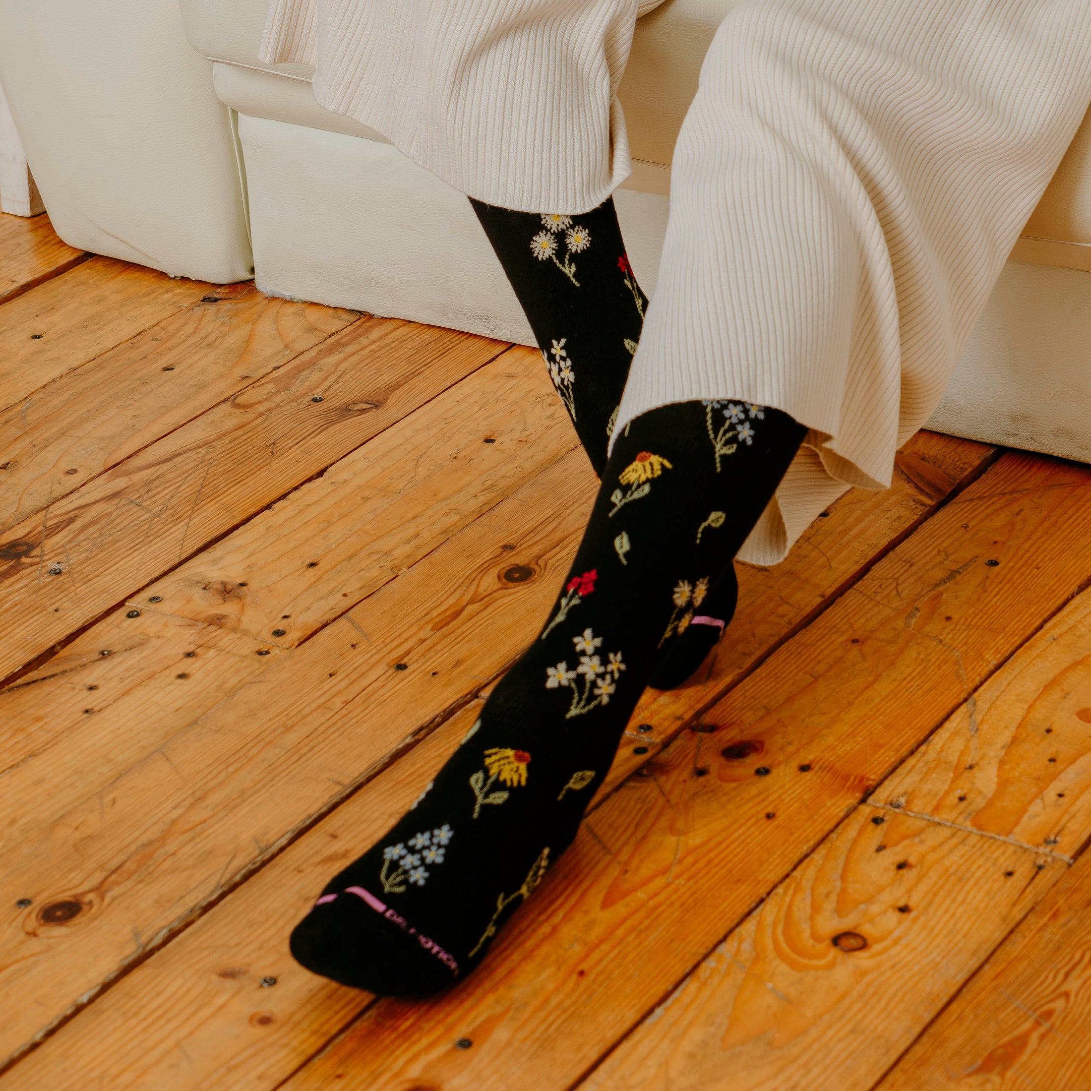 a cropped photo of someone in pajamas sitting on a couch wearing black sock printed with various sprigs of wildflowers