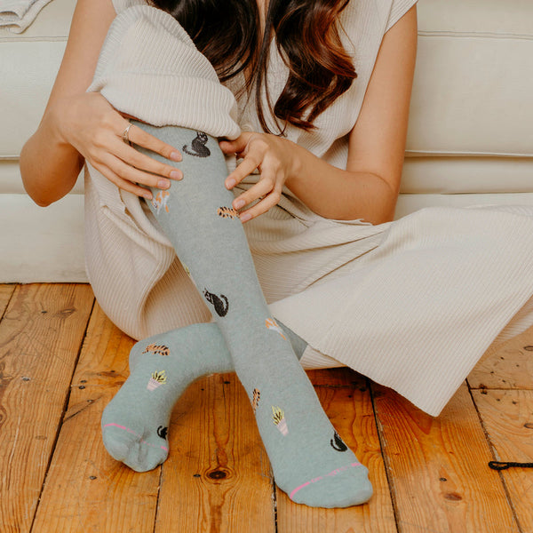 a woman seen from the shoulders down sitting on a wood floor wearing pajamas and light sage green heather socks featuring small cats in various colors and little potted plants