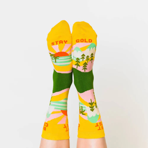 A model wearing yellow cotton women's novelty crew socks by the brand Yellow Owl Workshop feature green trees, teal and white mountains, and pink clouds and say the words 