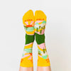 A model wearing yellow cotton women's novelty crew socks by the brand Yellow Owl Workshop feature green trees, teal and white mountains, and pink clouds and say the words "West Coast Best Coast" near the cuff and "Stay Gold" by the toes.