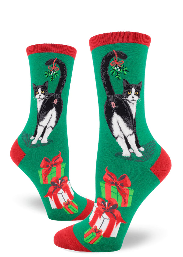 Shown on leg forms, a pair of women's crew sock sin green with a red heel, toe, and, cuff. The socks feature a tuxedo cat showing their butthole with a mistletoe hanging on the tail. The foot of the socks has Christmas presents in white, red, and, green.