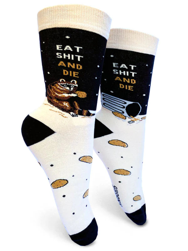 A white background shows a pair of crew socks on foot forms. The feet of the socks are beige with black toes & heels and a pattern of small black dots and large chocolate chip cookies. The ankle is black with several brown and white dots that look like stars. On the outer edge of the ankle a pudgy raccoon sits on its haunches with a cookie under beige and brown text that reads 