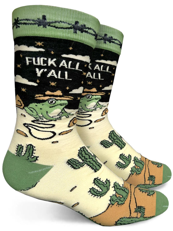 White background with two  black crew socks on foot forms with green barbed wire pattern around ankle, black background, beige and tan sand on foot, multiple green cactuses, and a green frog wearing a tan cowboy hat holding a whip. Beige text reads "FUCK ALL YALL."