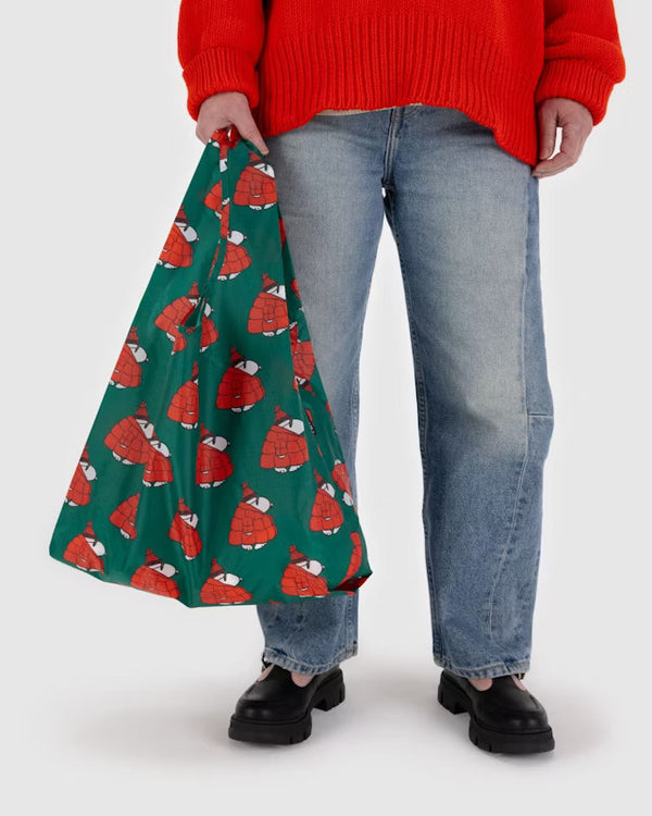 A model shown from the waist down holds a forest green canvas bag with several Snoopys walking as they wear sunglasses, red beanies and large red puffer coats.