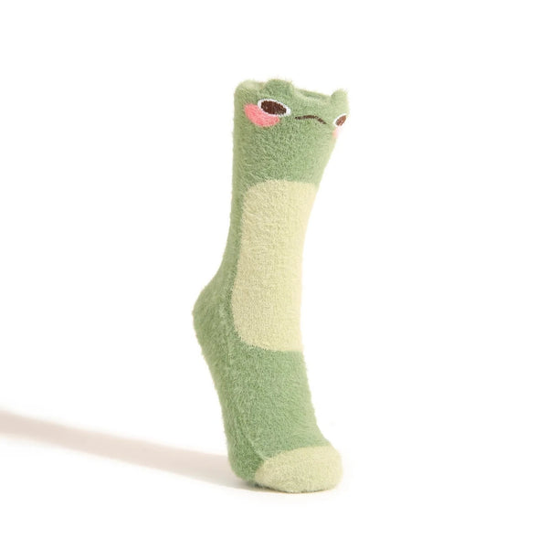 A green fuzzy crew sock with a blushing frog face at the top is shown on a foot form against a white backdrop.