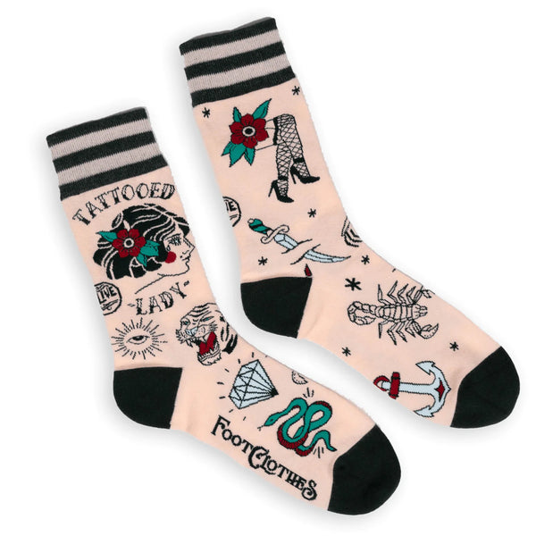 Shown in a flatlay, a pair of unisex crew socks in light pink with a gray heel and toe. These socks feature an all over motif of classic tattoo art with the words "Tattooed Lady" on the leg.